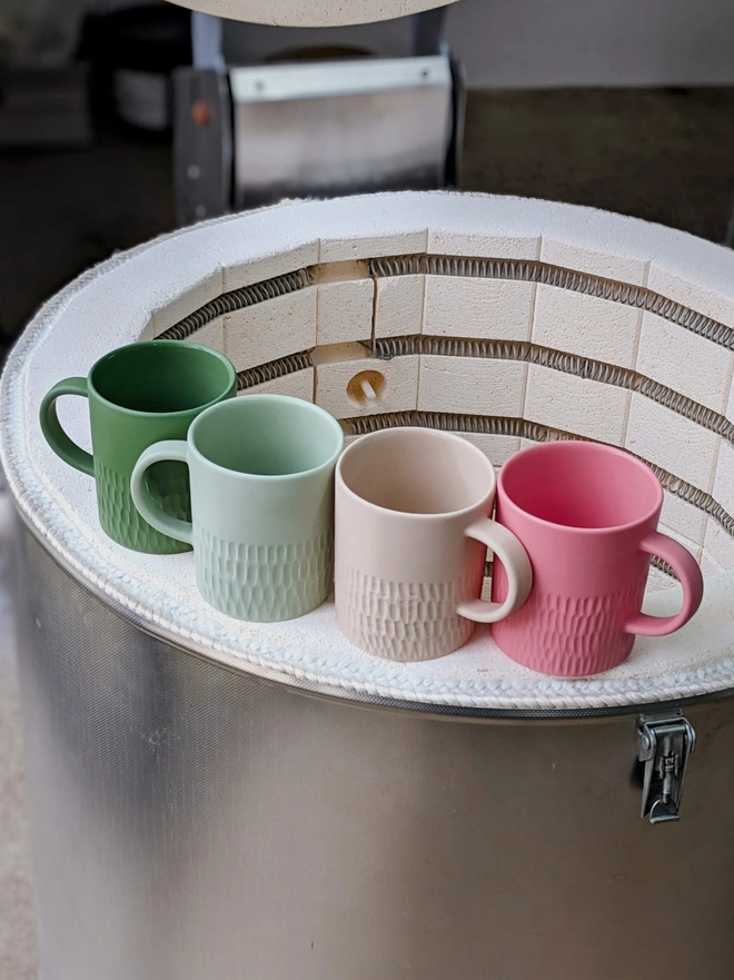 Four Large Mugs in Angelica Dark Green, Rockpool Pale Green, Neutral Pale Oat and Milkshake Pink sat on the edge of a kiln in pottery studio