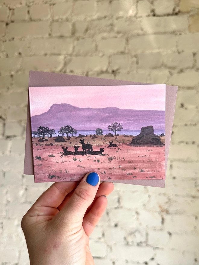 a greetings card featuring an illustration of a dusky coloured landscape and the silhouettes of some african wild dogs with a purple envelope