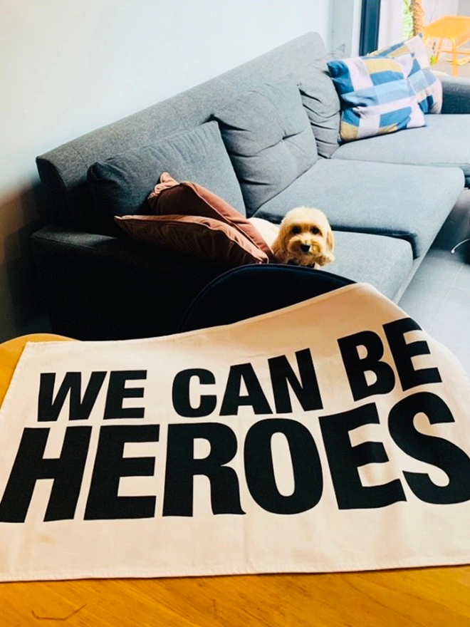 We Can Be Heroes printed in black on white tea towel laying on table with dog on sofa in background