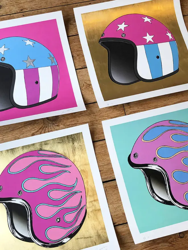 Four square art prints of motorcycle helmets. Two show a star-and-stripe design on pink and gold backgrounds; two show a flame design on gold and teal backgrounds.
