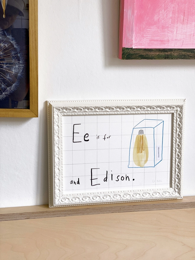 framed drawing of Edison lightbulb and hand written E for Edison in white frame on shelf 