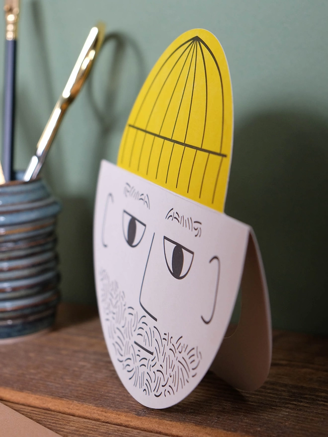 Side view of a round card with a male bearded face and yellow pop up hat. It is standing on a wooden shelf with green background.