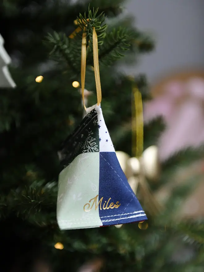 A handmade fabric fortune cookie Christmas decoration in a traditional patchwork print, personalised with the name “Miles” in gold, hangs from a golden ribbon on a Christmas tree branch.