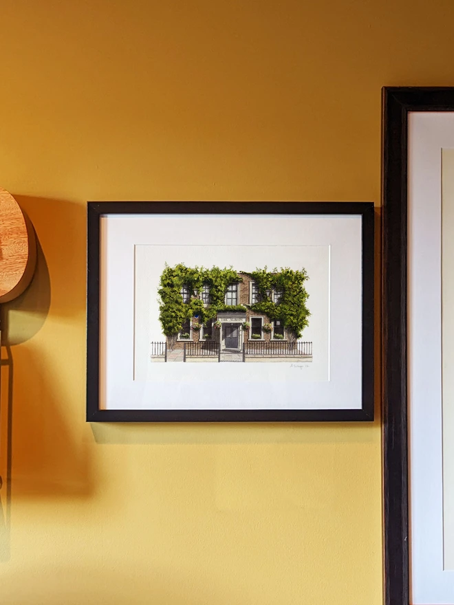 A framed watercolor print of The Albion hanging on a yellow wall next to a ukulele and another framed artwork.
