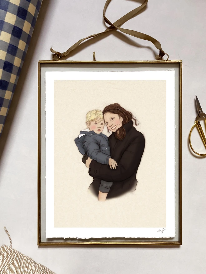 a photo of a personalised drawing of a mother holding her toddler framed in a glass frame with a brass edge and a velvet ribbon 