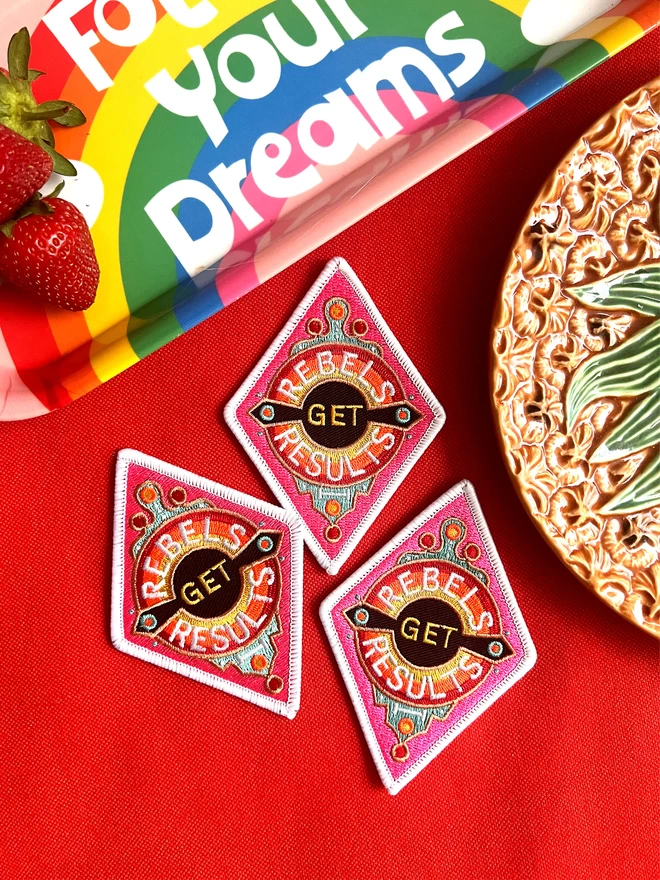 Three patches sit on a red table cloth next to a rainbow tray saying “follow your dreams” and part of an abstract pineapple ornament. The patch is a diamond shape embroidered patch with “Rebels Get Results” at the centre. The design features pink, white, red, yellow and black colours and has a white border.