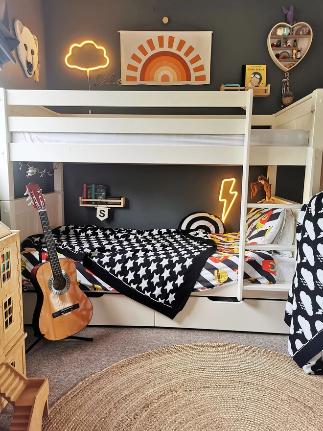 A view of white single bunk beds in a children's room, a black and white blanket with a star pattern is draped at an angle over the bottom bunk. Above the bed hangs a fabric banner with an orange sunset, and cloud and lightning bolt neon wall lights are switched on creating a warm and cosy feel. A guitar is propped on a stand near the side of the bed, and in the foreground a wooden dolls house is just visible.