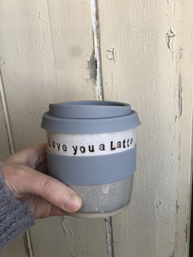 Personalised Short Ceramic Travel Mug, a blue ceramic travel mug sitting being held by a person in front of a wooden background. It's personalisation reads: ' Love you a latte'. 