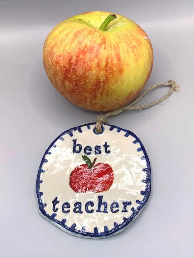 'Best Teacher' Handcrafted Ceramic Ornament, a ceramic apple ornament sitting on a plain white surface joined by an apple. 