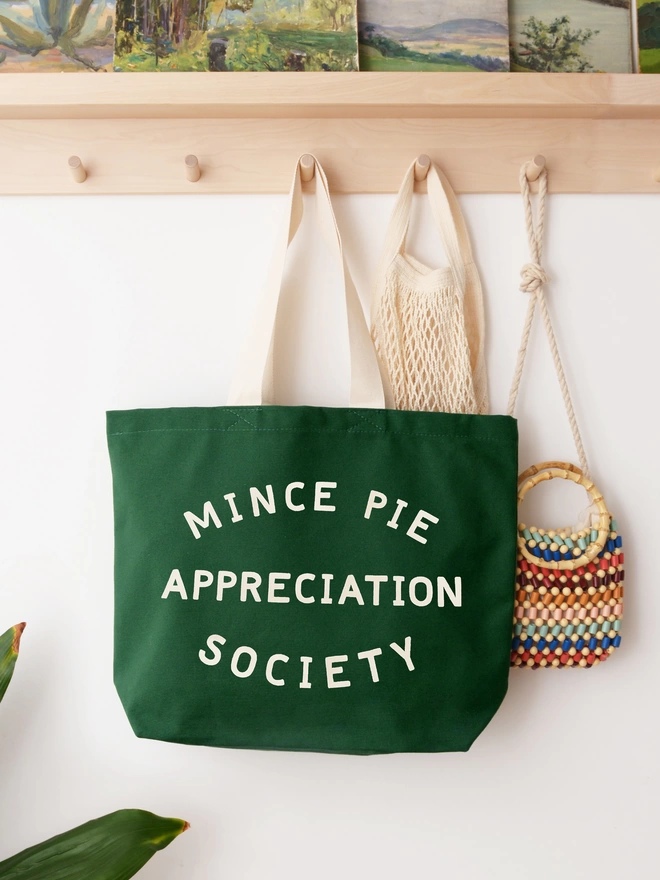 Green canvas Mince Pie Appreciation Society tote bag hanging from wooden hooks