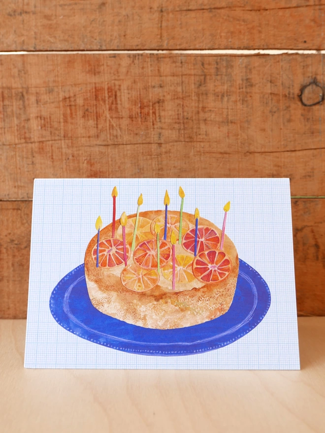 An illustration of an Orange Upside Down Birthday Cake on blue plate on a landscape format card
