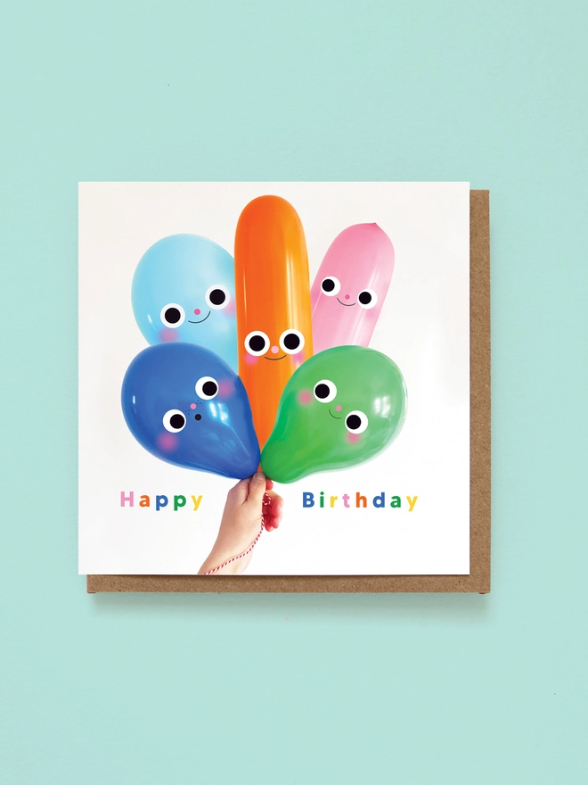 A birthday card with a hand holding a big bunch of colourful balloons with faces on. The card reads happy birthday 