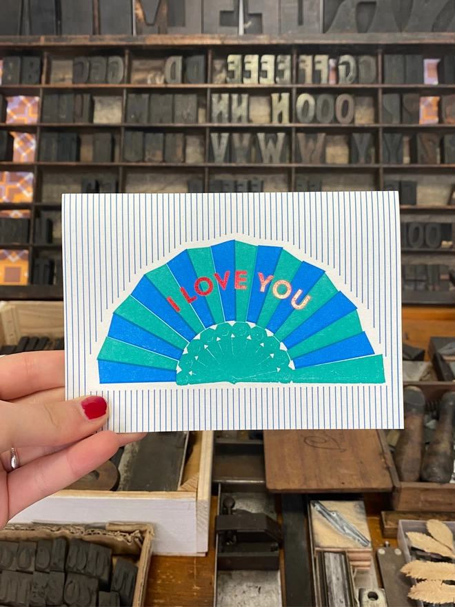 A hand holds up a white card with blue and green fan with red text reading 'I Love You' on green and white striped card.