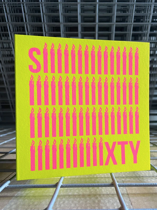 The word Sixty screen printed in neon pink ink across a bright yellow square card. To emphasise the sixty, the letter i is shaped like a simple lit candle and is repeated sixty times in four rows. The card is stood on a slight angle on a drying rack.ey studio set