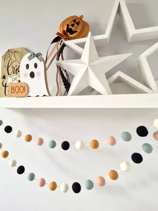 A white shelf filled with Halloween decor. Underneath is hanging a felt ball garland in peach, duck egg, ochre, ivory and black.