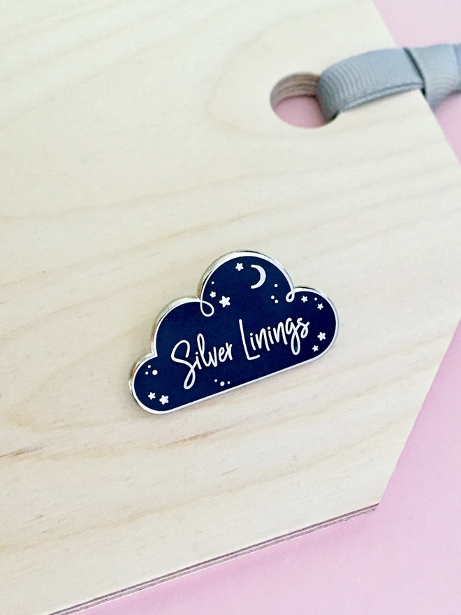 A navy blue and silver enamel pin badge in the shape of a cloud with a starry design and the words "Silver Linings" is placed on a wooden surface.