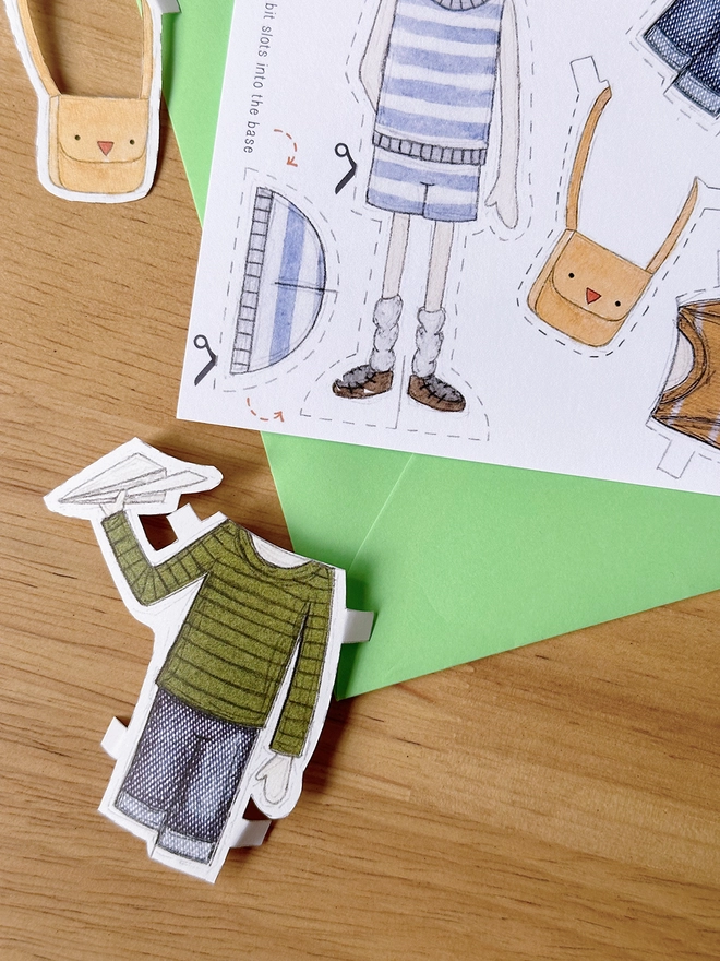A paper doll greetings card with a paper doll and several outfits on the card lays on a green envelope on a wooden desk.
