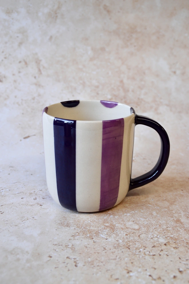 Handmade purple and cream striped ceramic mug with dark plum handle, photographed on a neutral stone background.