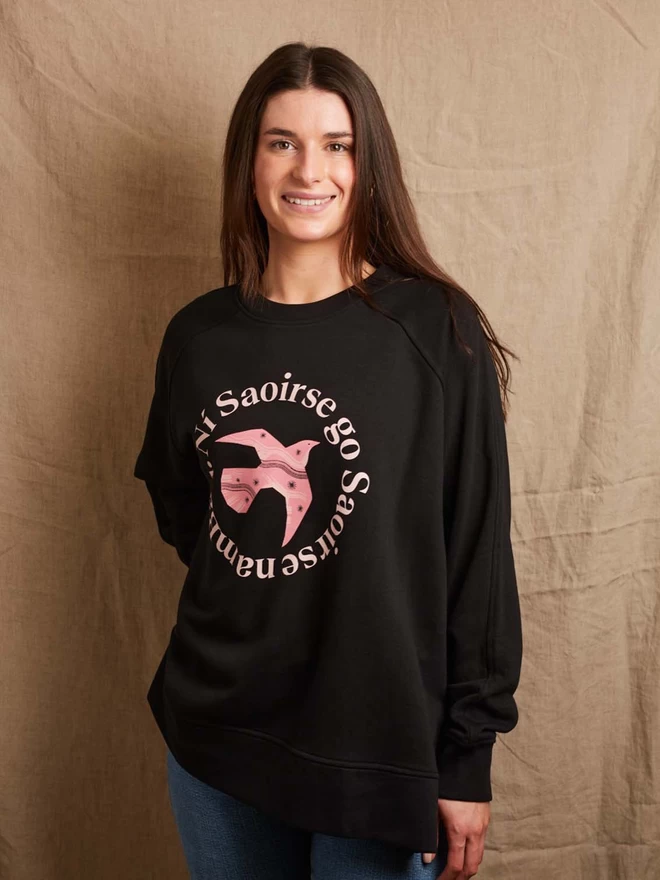 Model is wearing a boxy black sweatshirt with an image of a pink bird in flight, in the centre surrounded by the motto Ní Saoirse go Saoirse na mBan 