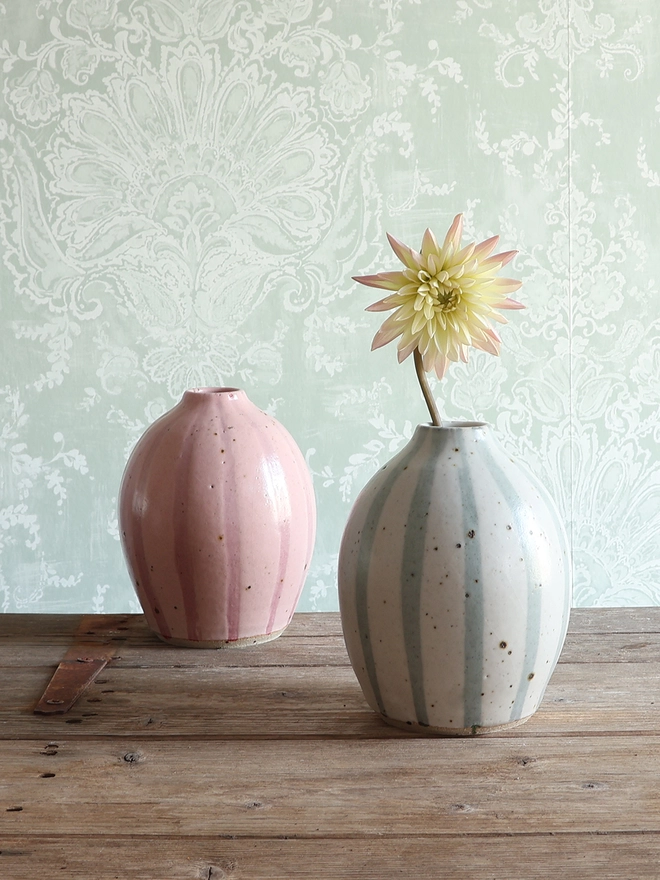 Handmade Ceramic Stoneware Rounded Vase in Blue and White Stripe holding a yellow and pink flower stem. The vase sits on a wooden surface with another vase in the same shape with a pink stripe design sat behind it.