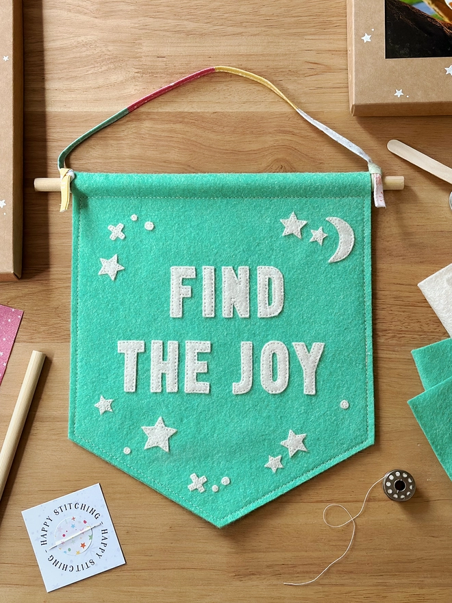 A turquoise felt banner with white words that read "Find The Joy" stitched on lays on a wooden desk.