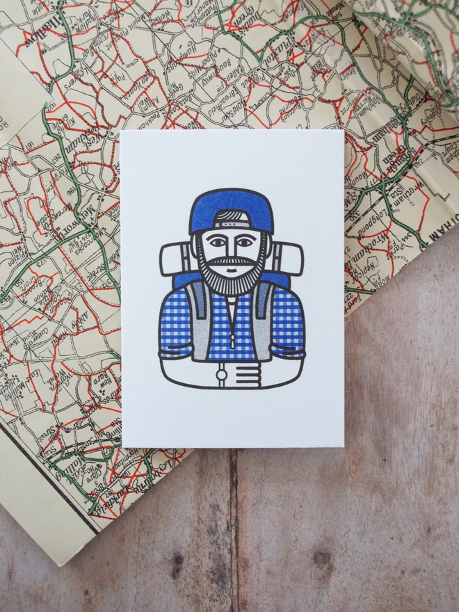 A bold modern illustration of a classic hiker in a blue gingham shirt with cap, rucksack and watch is printed onto a mini greeting card and placed on a vintage map.