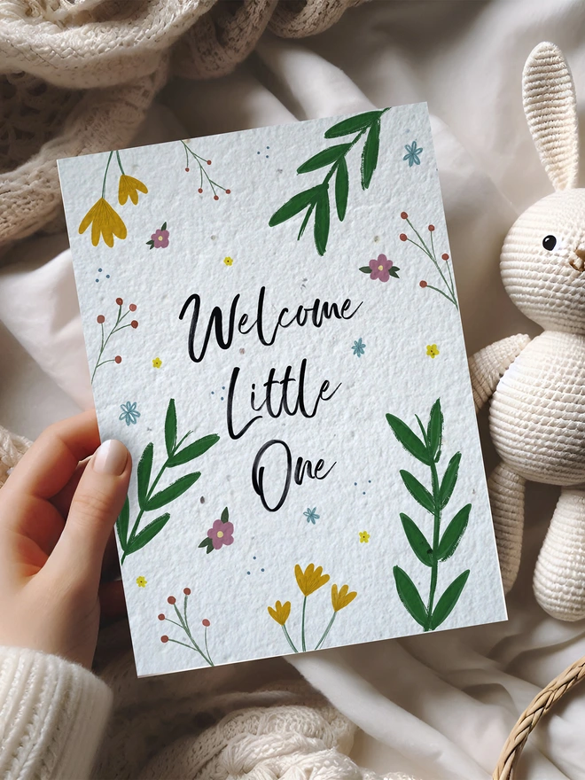 Hand holding a card with 'Welcome Little One' and a floral border. The card is placed on soft throws and a toy rabbit