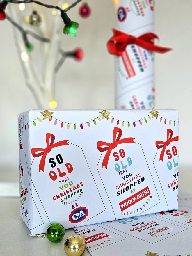 A photograph of a present wrapped in paper that has tags with a red bow printed on it. On each tag says so old that you Christmas shopped at Woolworths, Athena, C&A, Tie Rack, Sock Shop illustrated using brightly coloured text and the retro shop logo.