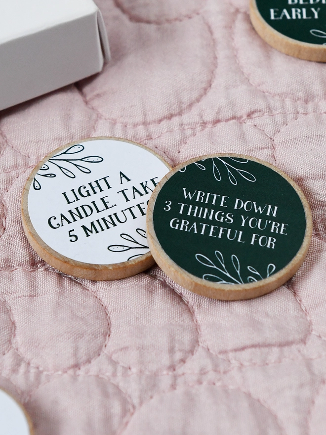 Two wooden tokens, each with a self care idea on, lay on a pink quilt.