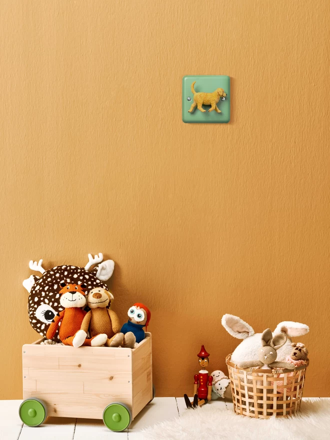 A pastel green nursery dimmer light switch with a puppy dog as the rotary knob sits on a beige wall. The light switch plate is beryl green and made of metal, epoxy coated steel. The puppy dog is made of plastic. The brand is Candy Queen Designs. On the floor there is a wooden toy boy with green wheels filled with soft toys and a wicker basket filled with soft toys too. The floor has a soft fluffy white rug on white wooden floor boards.