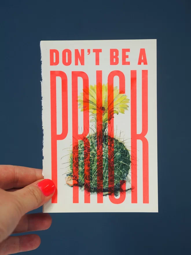 Hand holding a hand pulled screen print over a vintage book page of a cactus with the words ' don't be a prick' over the top in flouro red ink