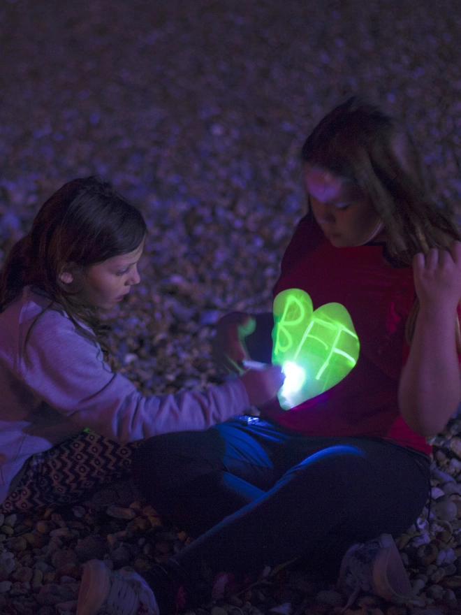 Girls on beach with one drawing with a torch onto another girls glow in the dark heart tshirt
