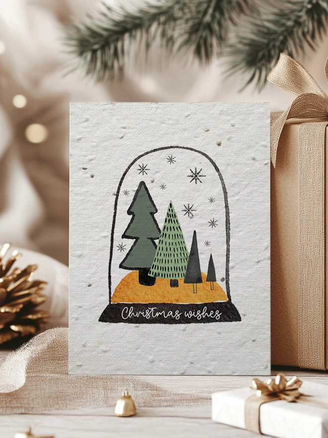 Christmas card with an illustrated snow globe on it with trees, snowflakes and 'Christmas Wishes'. The card is surrounded by presents, a pine cone and pine leaves.