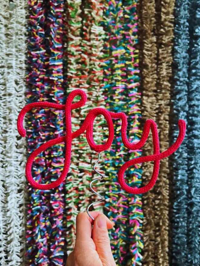 A hand dyed red cotton string tree topper spelling out the word 'Joy' held up by hand in front of a display of multicoloured Strinsel