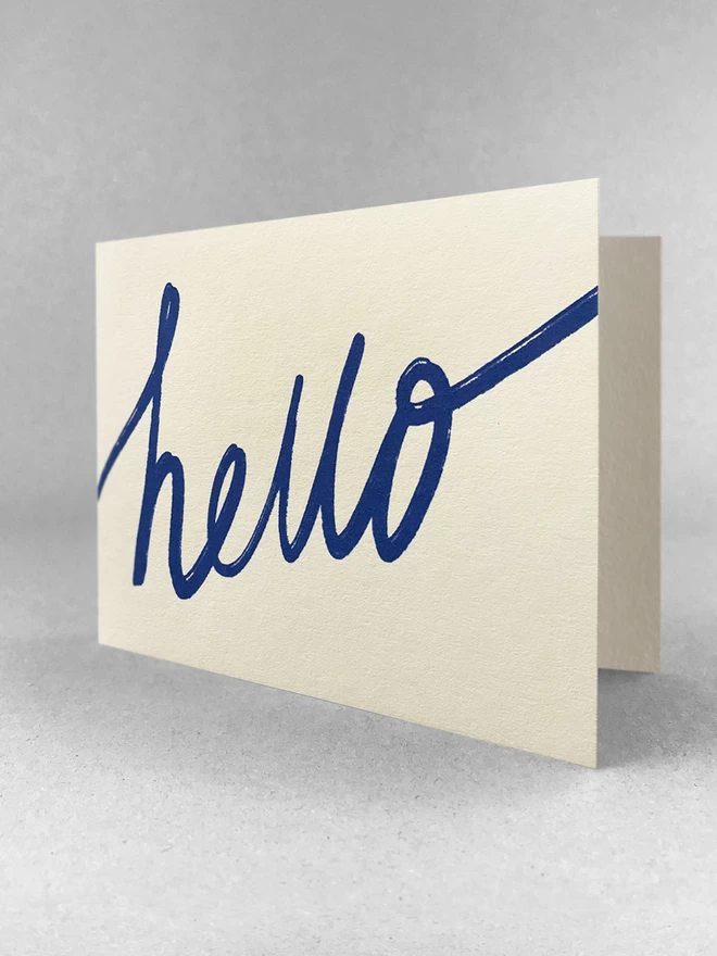 Handwritten blue hello is screenprinted on this landscape off-white greetings card. Stood slightly open, in a light grey studio set.
