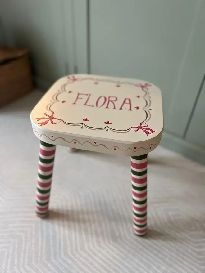 personalised hand-painted nursery stool. a small white stool with red and blue striped legs, painted with 'flora' on top.