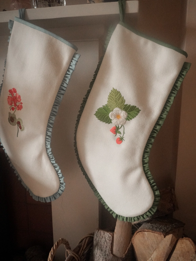 Two handmade ivory Christmas stockings hanging from a fireplace, embroidered with colourful floral and strawberry designs and finished with gathered fabric trims.