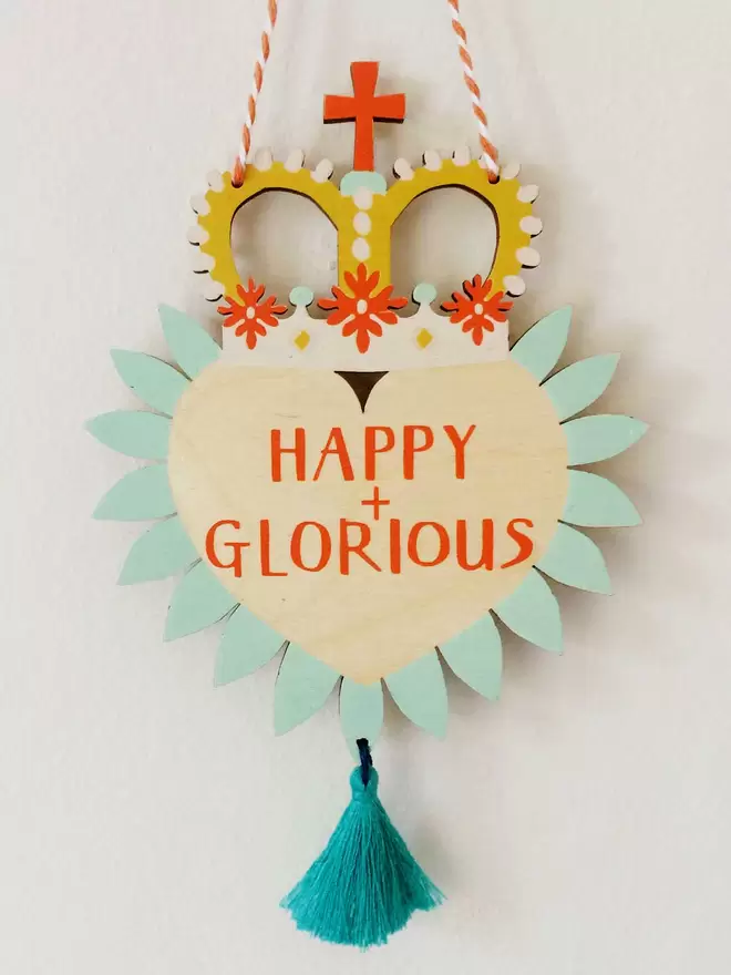 'Happy + Glorious' is hand printed on a colourful wooden heart with party crown and tassel. 