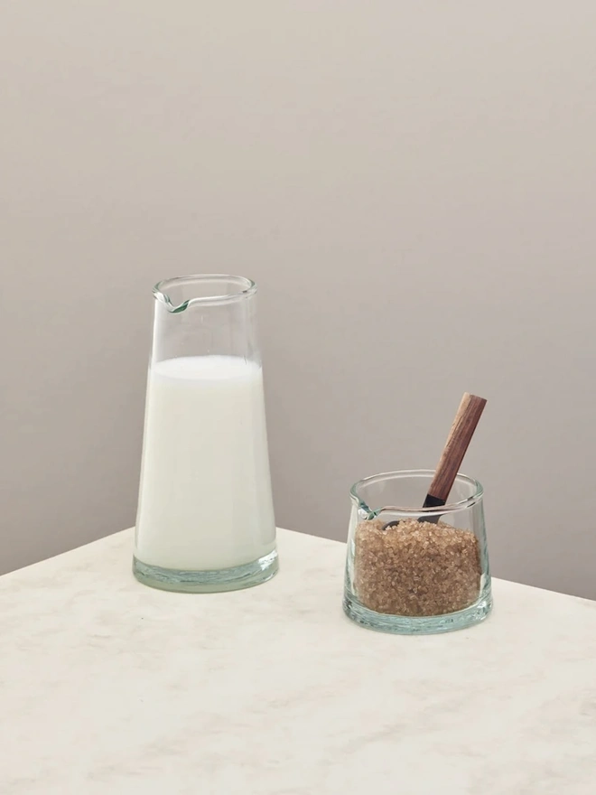 Glass Jugs (Set Of 2), a pair of a large and small glass jar sitting on a plain counter top against a grey backdrop. One is filled with sugar, the other with milk. 