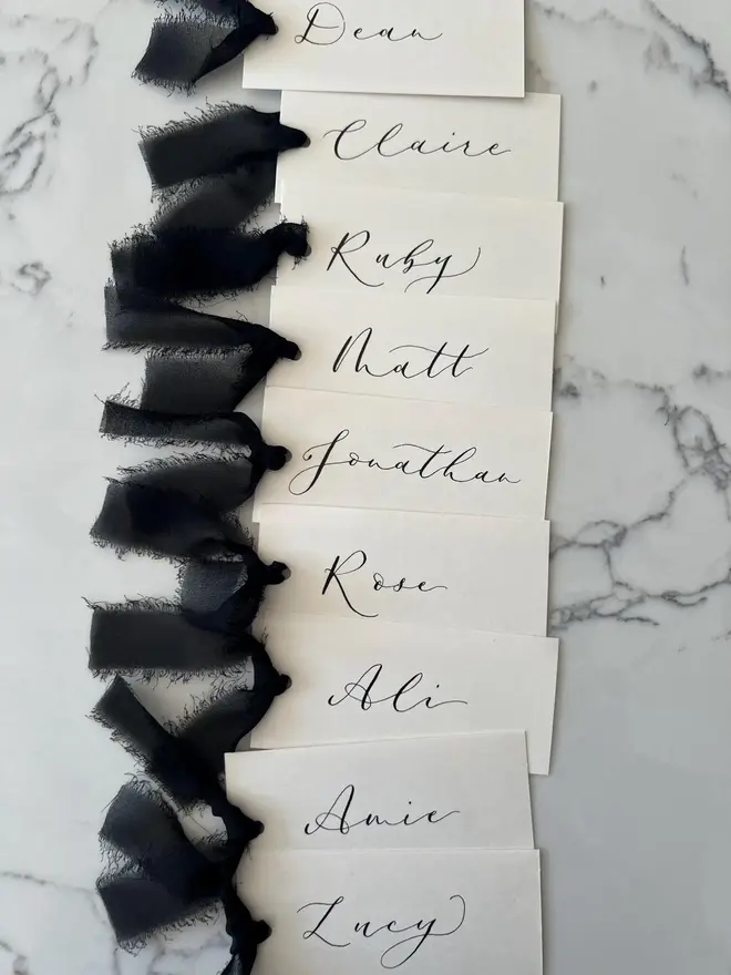 Personalised Calligraphy Wedding Cream Place Cards, cream place cards set together on a marble surface with black ribbons 