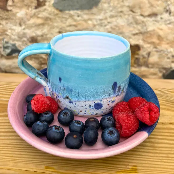 Shoreline Large Round Mug, a colourful mug on a wooden surface against a brick backdrop. It is accompanied by fruits. 