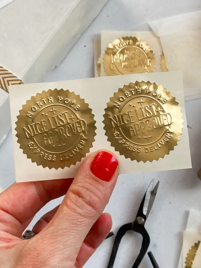 Two embossed gold "Nice List Approved" seal stickers on a cream backing sheet, held up by hand.