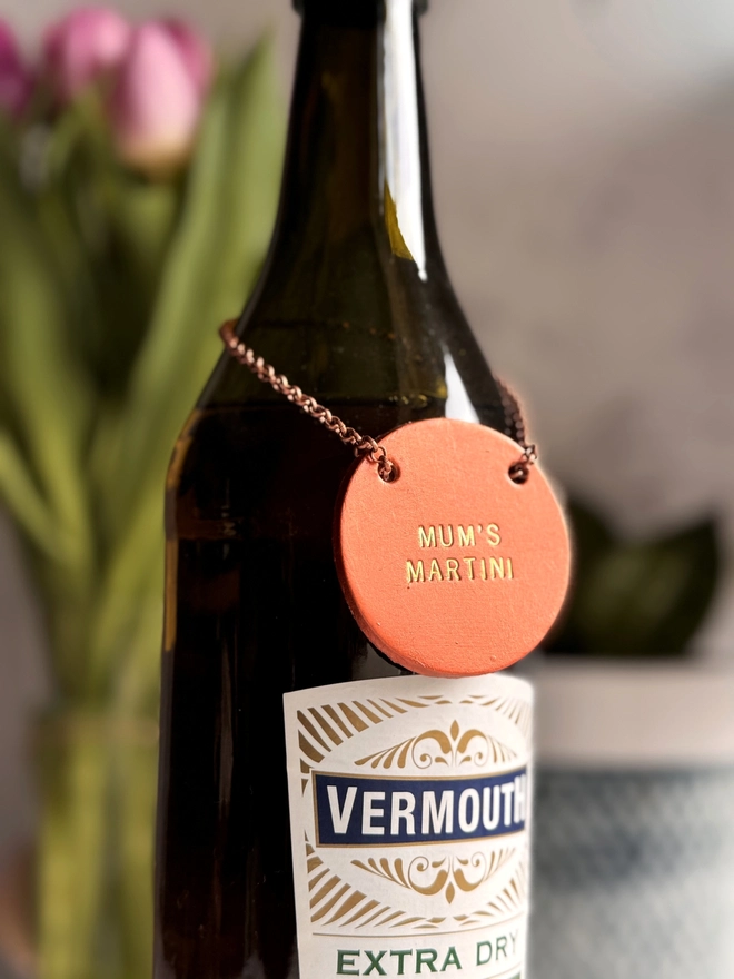 A dark glass bottle with a copper leather tag hanging around the neck by a delicate chain. The tag is hand-stamped with 'MUM'S MARTINI' in gold lettering, with pink tulips and a rustic background. A perfect personalised Mother’s Day gift or home bar accessory