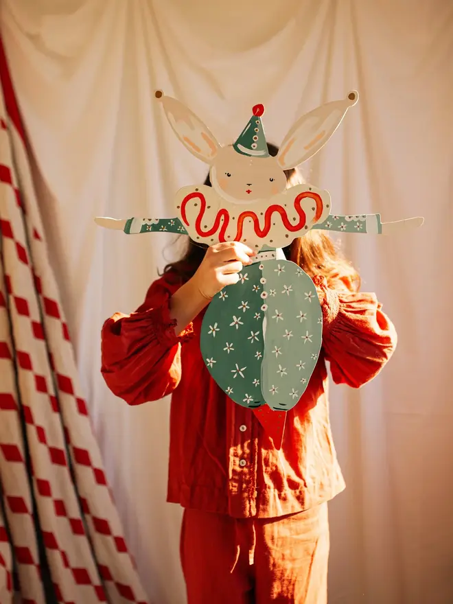 Extra Large Bella Hare Wall Puppet. a girl in red pyjamas holds the extra large bella hare wall puppet up in front of her face against a red-and-white striped curtain and cream backdrop. the hare wears a white harlequin-style outfit with a swirling orange motif, teal floral leggings, and a small pointed party hat, with jointed limbs painted in soft cream and teal tones. the warm, golden light and playful pose give the puppet a nostalgic, storybook charm.
