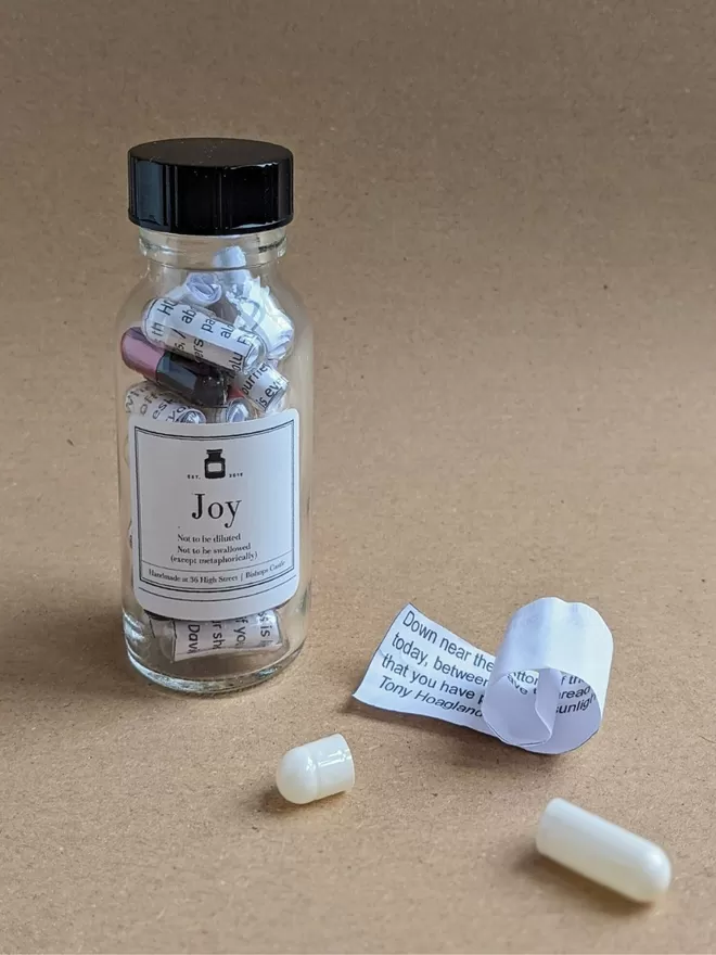 Glass bottle containing black and rose gold Joy poetry pills printed on banana paper