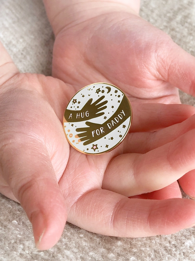 A child is holding a white and gold pin badge in their hands. It has two arms in a hugging design with the words "A hug for Daddy".