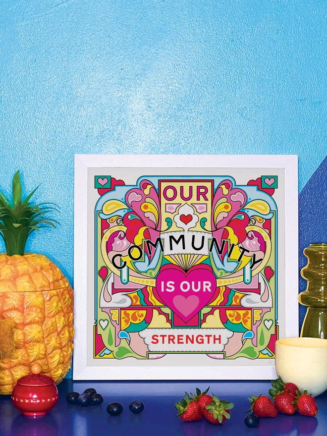 Our Community is Our Strength is written over this bold, symmetrical illustration of yellows, greens and pinks. It is in a white square frame propped against a wall painted light and dark blue, on a shiny blue cabinet. Next to the frame is a plastic pineapple ice bucket, a small wooden red pot a handful of blueberries, a yellow glass vase, a small yellow ceramic pot and 4 strawberries.