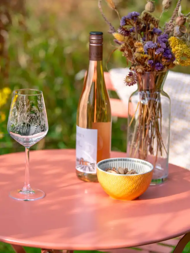 Meadow Wine Glass, an etched wine glass with a wildflower pattern sits on a bright pink patio table next to a bottle of rosé wine.