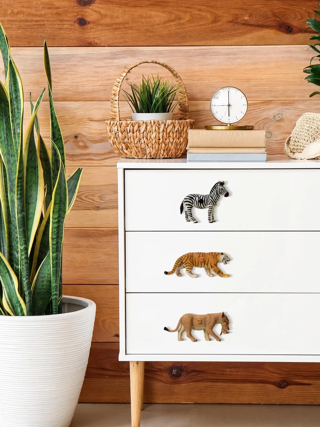 A white chest of drawers with wooden legs against a medium brown pine clad wall. On the drawers are three safari animal knobs made by Candy Queen Designs. On the top drawer is a zebra drawer knob, on the middle drawer there is a tiger drawer knob and on the bottom drawer there is a lioness drawer knob. The animal knobs are made of plastic. To the left of the drawers is a snake plant in a big white plant pot. On top of the drawers there is a small plant in a wicker basket, a small clock and three books with plain covers.