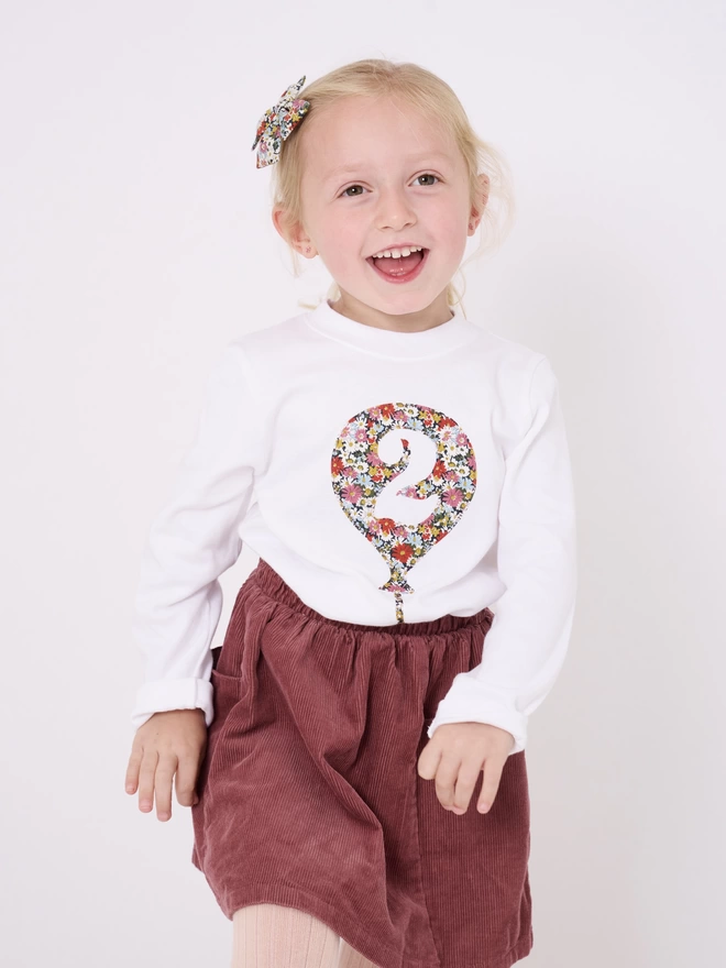 A smiling 2 year old girl wearing a white cotton long sleeve t-shirt. The t-shirt features a balloon with the number 2 cut out from it, appliquéd in a floral Liberty print.